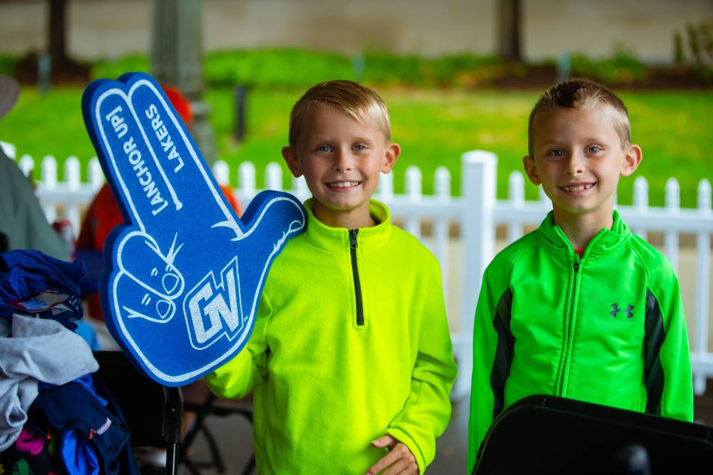 Two younger siblings at the gvsu party are standing and smiling for a photo. One is holding up his gvsu foam finger